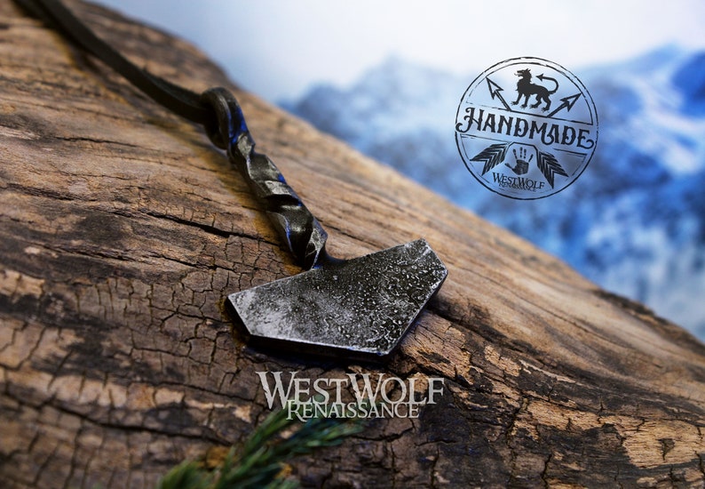 May include: A silver-colored metal hammer pendant with a twisted handle, hanging from a black leather cord. The pendant is on a wooden surface with the text "WESTWOLF RENAISSANCE" below it. A circular logo with the text "HANDMADE" and a wolf design is visible in the background.