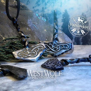 May include: Two silver-colored Mjolnir pendants with intricate knotwork designs. The pendants are on a black leather cord and a twisted metal chain. The background is a snowy landscape with a handmade logo.