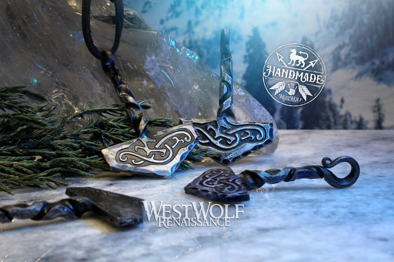 May include: Two silver-colored Mjolnir pendants with intricate knotwork designs. The pendants are on a black leather cord and a twisted metal chain. The background is a snowy landscape with a handmade logo.