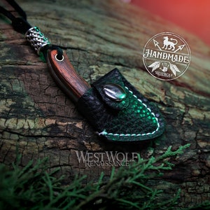 May include: A small, handmade knife with a wooden handle and a leather sheath. The knife is attached to a black cord and has a silver bead at the top. The knife is resting on a piece of wood with the text "WESTWOLF RENAISSANCE" visible below it.