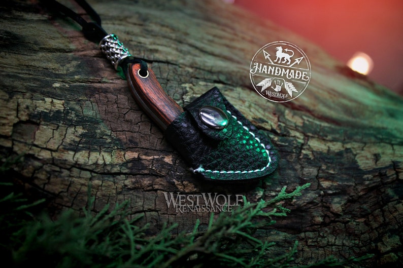 May include: A small, handmade knife with a wooden handle and a leather sheath. The knife is attached to a black cord and has a silver bead at the top. The knife is resting on a piece of wood with the text "WESTWOLF RENAISSANCE" visible below it.