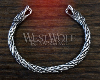 May include: Silver bracelet with a braided design and two wolf head ends. The bracelet is on a wooden surface.