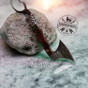 May include: A small, silver axe pendant with a wood handle and a leather cord. The axe has a decorative, wavy pattern on the blade. The pendant is resting on a green rock with a silver bead on the cord. The image includes the text "Handmade" and "Westwolf Renaissance".