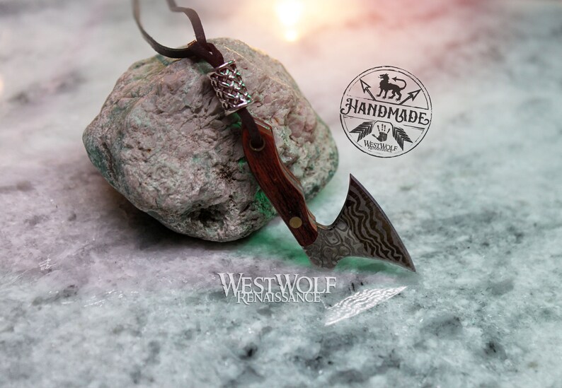 May include: A small, silver axe pendant with a wood handle and a leather cord. The axe has a decorative, wavy pattern on the blade. The pendant is resting on a green rock with a silver bead on the cord. The image includes the text "Handmade" and "Westwolf Renaissance".