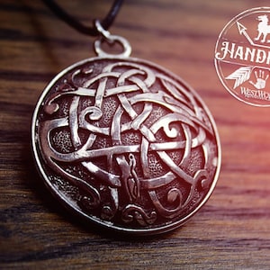 May include: Silver Celtic knot pendant necklace with a detailed intricate design. The pendant is round and has a textured surface. The necklace is on a black cord.