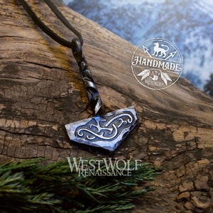 May include: A silver and black metal pendant in the shape of a hammer with intricate designs. The pendant is hanging from a black leather cord. The pendant is on a wooden surface with a blurred background of snow-covered mountains.