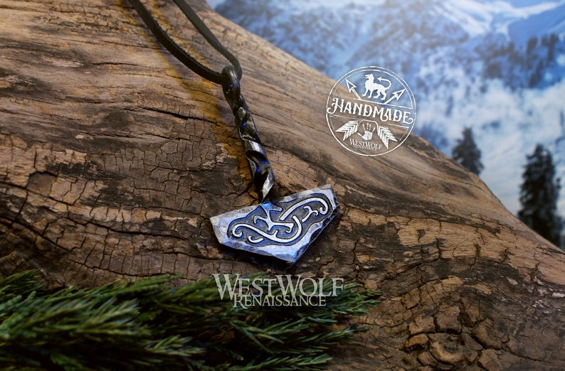 May include: A silver and black metal pendant in the shape of a hammer with intricate designs. The pendant is hanging from a black leather cord. The pendant is on a wooden surface with a blurred background of snow-covered mountains.