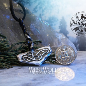 May include: A silver-colored metal pendant in the shape of a hammer with intricate designs. The pendant is hanging from a black leather cord. The pendant is next to a US quarter for scale. The image includes the text "Westwolf Renaissance" and "Handmade Westwolf Renaissance".