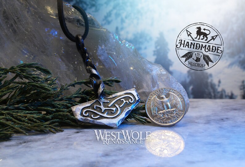 May include: A silver-colored metal pendant in the shape of a hammer with intricate designs. The pendant is hanging from a black leather cord. The pendant is next to a US quarter for scale. The image includes the text "Westwolf Renaissance" and "Handmade Westwolf Renaissance".