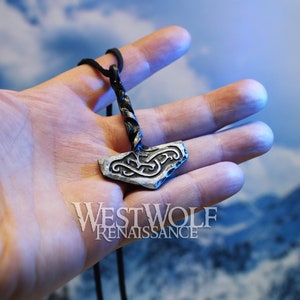 May include: A silver Mjolnir pendant with a black cord necklace. The pendant has a twisted cord and a detailed knot design. The pendant is held in a hand against a blue and white background.