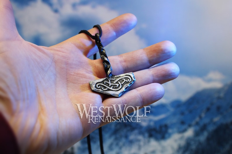 May include: A silver Mjolnir pendant with a black cord necklace. The pendant has a twisted cord and a detailed knot design. The pendant is held in a hand against a blue and white background.