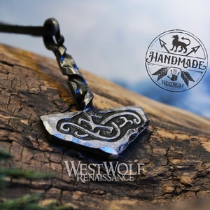 May include: A silver-colored metal pendant in the shape of a hammer with intricate knotwork designs. The pendant is attached to a black leather cord. The pendant is on a wooden surface. The image also includes a logo that says "Handmade Westwolf".
