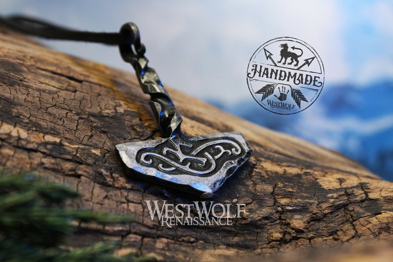 May include: A silver-colored metal pendant in the shape of a hammer with intricate knotwork designs. The pendant is attached to a black leather cord. The pendant is on a wooden surface. The image also includes a logo that says "Handmade Westwolf".