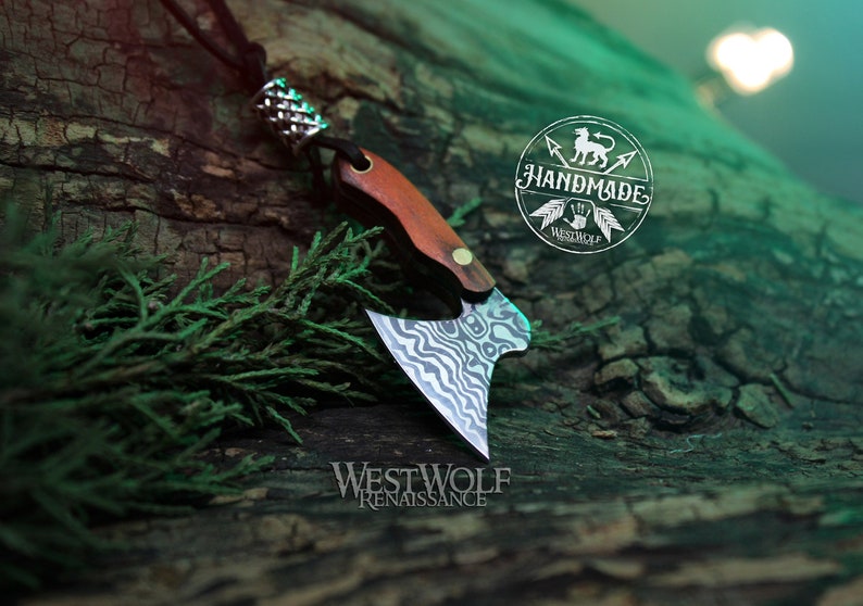 May include: A small, handmade axe pendant with a wood handle and a patterned steel blade. The pendant is on a black cord and is hanging from a tree branch. The pendant is labeled "Westwolf Renaissance" and "Handmade".