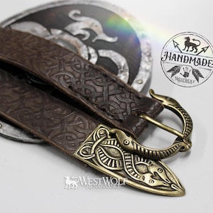 Puede incluir: Un cinturón de cuero marrón con una hebilla ornamentada con un diseño de animal estilizado. El cinturón tiene un patrón en relieve detallado. La hebilla y la punta del cinturón son de color dorado. La imagen también incluye un escudo y un logotipo "HANDMADE".