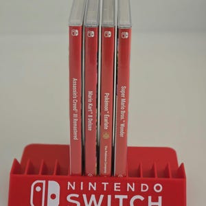 May include: A red plastic Nintendo Switch game storage stand holding four video game cases. The stand features the white Nintendo Switch logo. The game cases are Assassin's Creed III Remastered, Mario Kart 8 Deluxe, Pokémon Écarlate, and Super Mario Bros. Wonder.