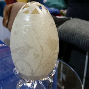 May include: A white ostrich egg with intricate carvings of flowers and leaves. The egg is displayed on a clear acrylic stand.