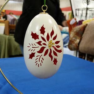 May include: A white egg-shaped ornament with a red poinsettia design and gold glitter accents. The ornament is hanging from a gold stand.