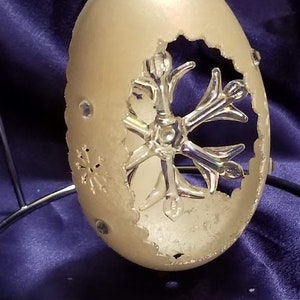 May include: A white, pearlescent, egg-shaped ornament with a cut-out snowflake design. The snowflake is made of iridescent glass and is attached to the ornament with a silver wire.