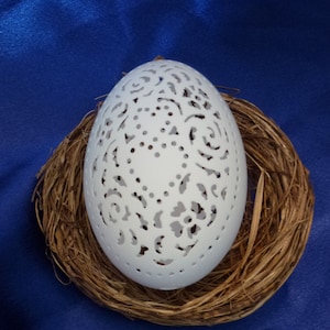 May include: A white egg with intricate floral designs carved into its surface. The egg is nestled in a brown woven nest.