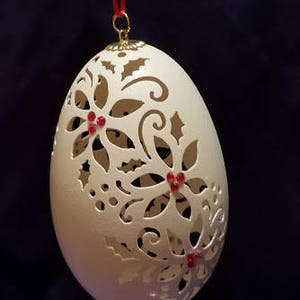 May include: A white, intricately carved egg ornament with a red ribbon and red jeweled accents. The egg is decorated with a floral design and holly leaves.