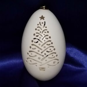May include: A white, intricately carved ostrich egg ornament with a Christmas tree design. The tree is carved with a star at the top and a gold-colored ornament at the bottom.