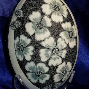 May include: A white ceramic egg with a black background and white flowers with green accents. The egg is on a clear plastic stand.