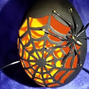 May include: Black egg-shaped ornament with a cut-out spider web design and a black spider silhouette. The web is illuminated from inside, creating a glowing orange effect.