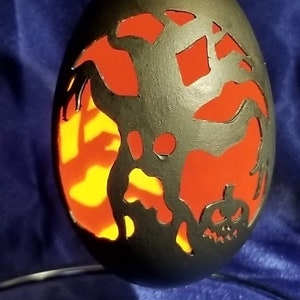 May include: A black egg-shaped ornament with a carved design of a ghost and a jack-o'-lantern. The carving is illuminated from behind, creating a glowing orange effect.
