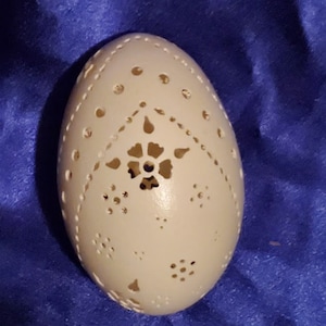 May include: A white egg with intricate carvings of a flower and geometric patterns. The egg is on a blue fabric background.