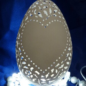 May include: A white, intricately carved egg with a delicate floral design. The egg is illuminated from below, casting a soft glow through the intricate carvings. The egg is displayed on a clear stand.