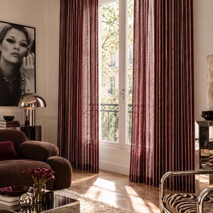 May include: Floor-to-ceiling curtains featuring a deep burgundy red fabric with thin, vertical white stripes. The curtains are hung on a rod above a window, with the fabric draping down to the floor, allowing light to filter through the material.