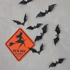 Pode incluir: Uma placa laranja em forma de diamante com borda preta e a silhueta preta de uma bruxa voando em uma vassoura. O texto diz BEWARE WITCHES CROSSING. Oito morcegos de papel preto estão dispostos ao redor da placa em várias posições.