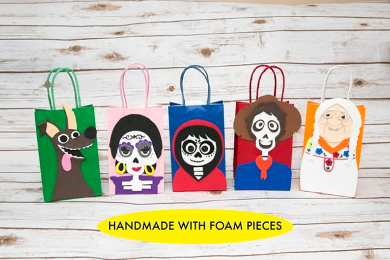 May include: Five colorful paper gift bags with different characters from the movie Coco. The bags are decorated with foam cutouts of characters like Miguel, Dante, and Mama Imelda. The bags are handmade and have the text "HANDMADE WITH FOAM PIECES" at the bottom.