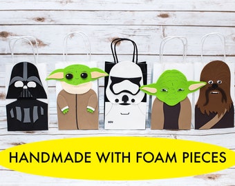 May include: Five white paper gift bags with different Star Wars characters made from foam pieces. The characters are Darth Vader, Yoda, Stormtrooper, Chewbacca, and Baby Yoda. The bags are handmade and have white handles. The text "HANDMADE WITH FOAM PIECES" is printed in yellow on a black oval.