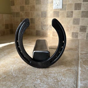 May include: A black horseshoe-shaped metal object with a rectangular metal bar attached. The horseshoe has visible nail holes and is positioned upright. The object is likely a decorative item or a functional piece for a home or stable.