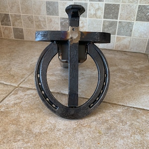 May include: A metal cross with a horseshoe attached to the bottom. The cross is made of black metal and has a hammered finish. The horseshoe is also black metal and has a rustic look.
