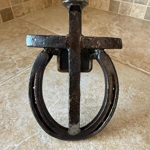 May include: A rustic, handcrafted metal cross featuring a horseshoe design. The cross is made of dark, weathered metal, with a vertical bar intersecting a horseshoe shape. The nail head at the top adds a unique detail to this decorative piece.