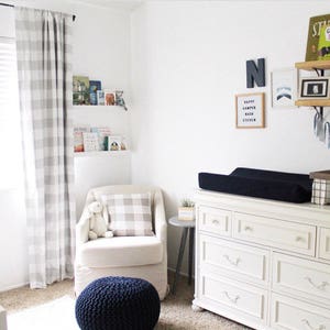 Puede incluir: Una c&oacute;moda blanca con seis cajones y una cambiador negro en la parte superior. La c&oacute;moda est&aacute; en una guarder&iacute;a con una silla mecedora blanca, una almohada de tiro de cuadros grises y blancos, un otomano azul y un estante de madera con libros y una guirnalda azul y blanca.