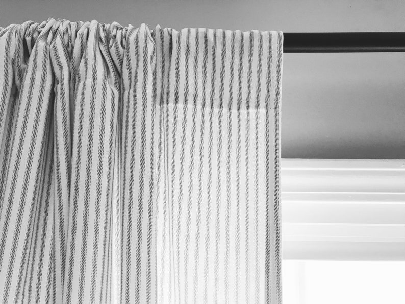 May include: A white curtain with thin black stripes hangs from a black curtain rod. The curtain is gathered at the top and the fabric is slightly wrinkled.