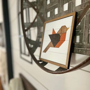 May include: A framed geometric art print of a bird with orange, brown, and black colors. The print is hanging on a white wall with a metal circular object behind it.