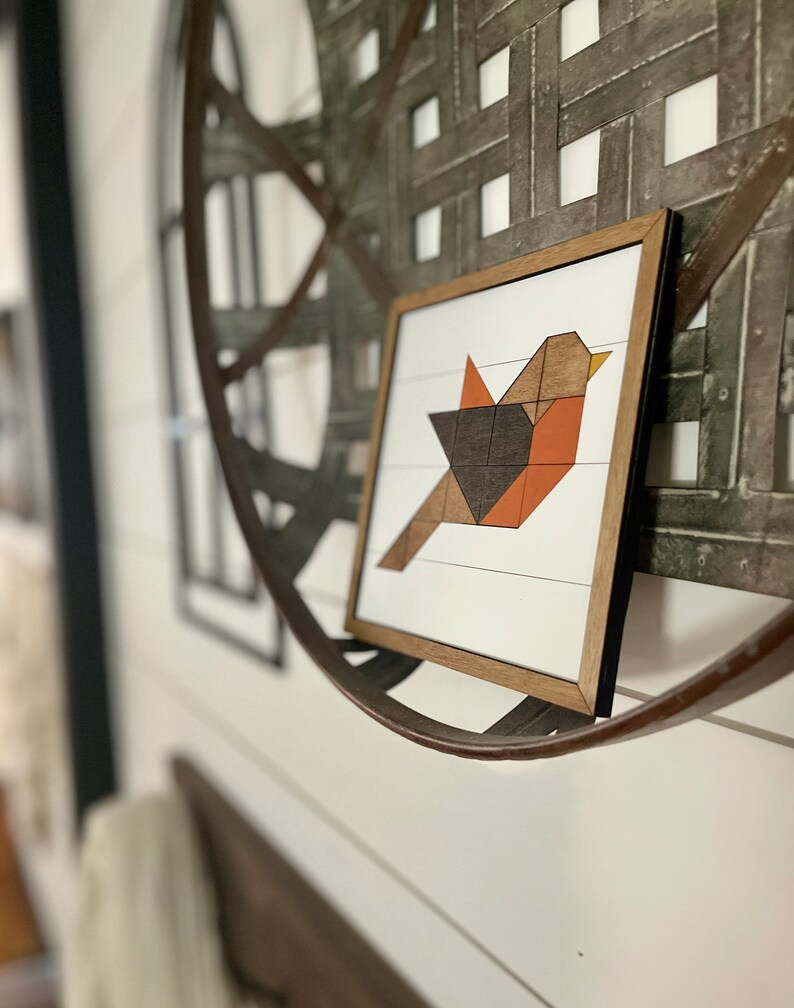 May include: A framed geometric art print of a bird with orange, brown, and black colors. The print is hanging on a white wall with a metal circular object behind it.