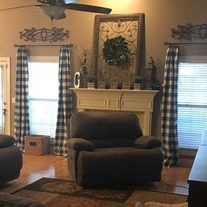 Puede incluir: Una sala de estar con una chimenea, dos sillones reclinables marrones y tres ventanas con cortinas a cuadros azules y blancas. Las ventanas tienen persianas blancas. Hay una estanter&iacute;a de madera en el lado derecho de la imagen.