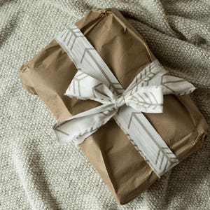 Puede incluir: Un paquete de papel marr&oacute;n envuelto con una cinta blanca con un patr&oacute;n geom&eacute;trico. La cinta est&aacute; atada en un lazo.