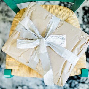 May include: A brown paper-wrapped gift box tied with a silver ribbon and a small white tag with black text. The gift is sitting on a woven chair.