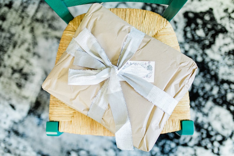 May include: A brown paper-wrapped gift box tied with a silver ribbon and a small white tag with black text. The gift is sitting on a woven chair.