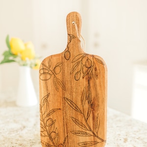 May include: A wooden chopping board with a natural wood finish and a handle. The board is decorated with a laser-etched design of olive branches and olives.