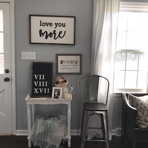 May include: A white side table with a blue blanket on the bottom shelf. The table has a black framed sign with Roman numerals VII, VIII, and XVIII. A white framed picture with a floral design is above the sign. A metal stool with a wooden seat is next to the table. The wall behind the table is painted a light gray color. A wooden sign with the text "love you more" is hanging on the wall. A white framed picture with the text "Little paws, tiny toes, little people, our family grows" is hanging on the wall above the sign.