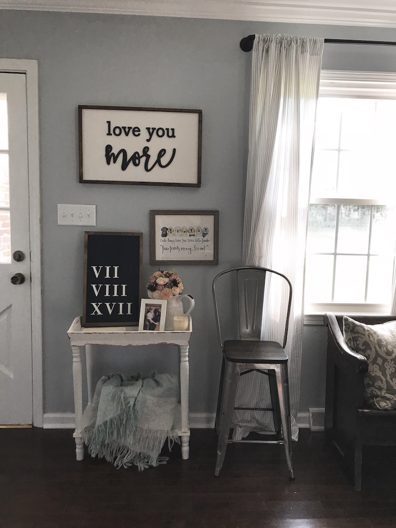 May include: A white side table with a blue blanket on the bottom shelf. The table has a black framed sign with Roman numerals VII, VIII, and XVIII. A white framed picture with a floral design is above the sign. A metal stool with a wooden seat is next to the table. The wall behind the table is painted a light gray color. A wooden sign with the text "love you more" is hanging on the wall. A white framed picture with the text "Little paws, tiny toes, little people, our family grows" is hanging on the wall above the sign.