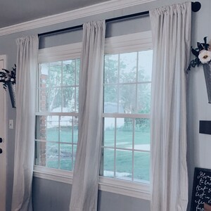 May include: Two windows with white striped curtains and a white door. The windows have a view of a green lawn and trees. There is a metal bucket with artificial flowers hanging on the wall to the right of the windows.